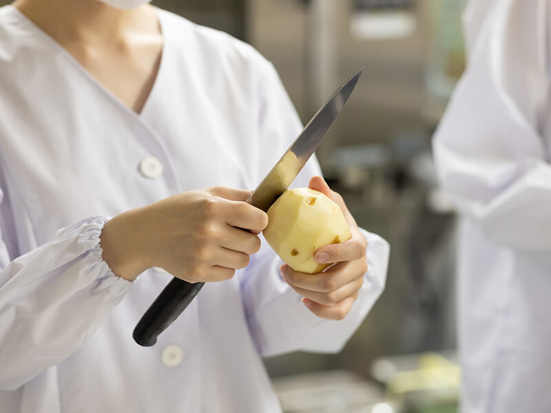 白衣を着た女性が包丁でジャガイモの皮を剥いているシーンです。