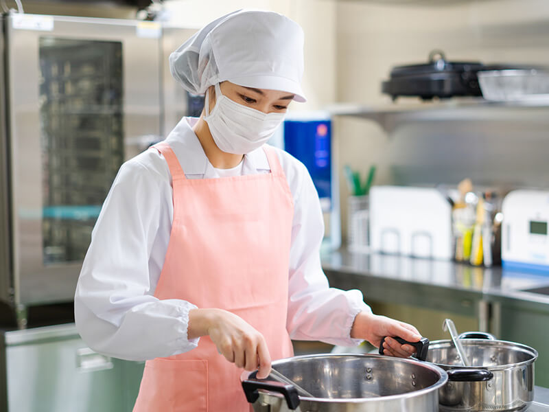 エプロンを着てマスクをした女性が厨房で料理をしている様子。